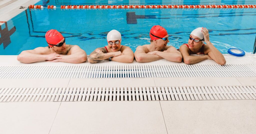 Four adult swimmers in caps and goggles relax by the indoor poolside, smiling and chatting.