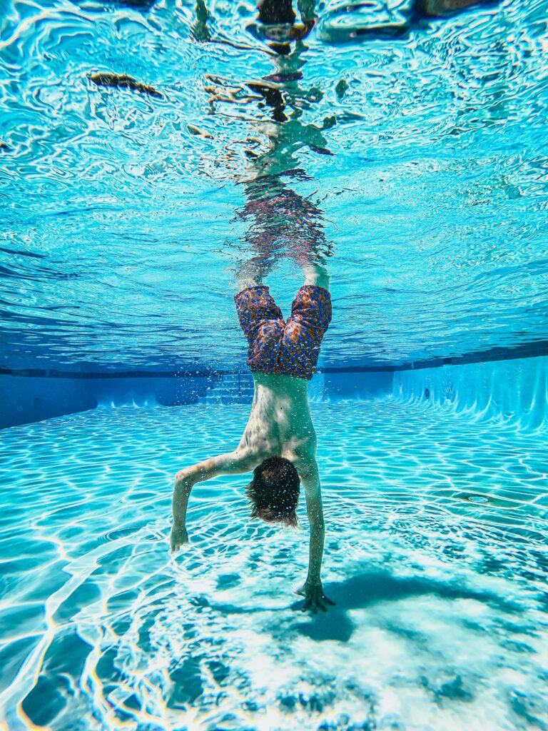 A child performs a handstand in a bright blue swimming pool, showcasing summer fun.