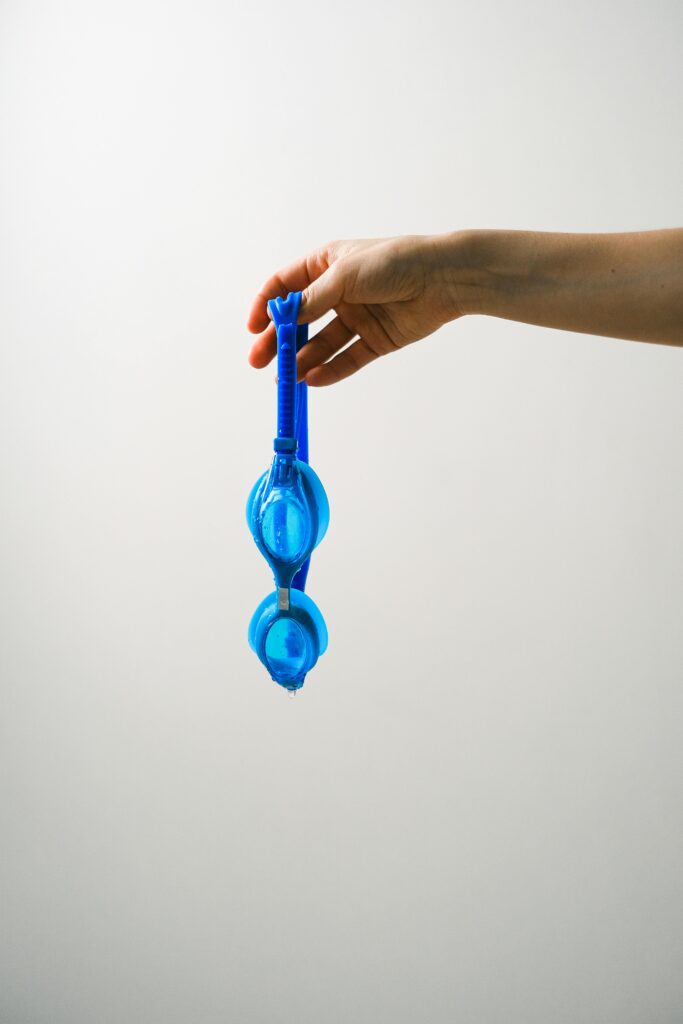 A close-up of a hand holding blue swimming goggles against a minimalist white background.